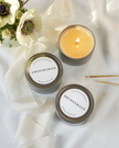 Two lit candles in tins labeled 'Groomsman' and 'Bridesmaid' on a white fabric background with flowers.