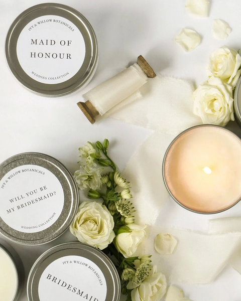 Candle with 'Maid of Honour' and 'Will you be my Bridesmaid?' tins surrounded by white roses on a white background.